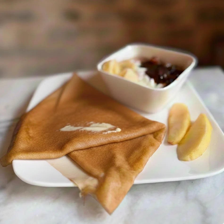 Une crêpe dorée pliée en triangle, nappée de beurre fondu, posée sur une assiette en porcelaine blanche décorée de baies, sur une table en bois texturé.