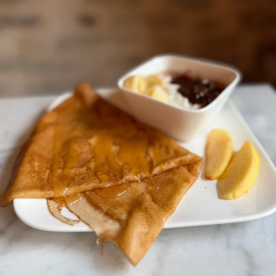 Une crêpe dorée pliée en triangle, généreusement nappée de miel liquide brillant, posée sur une assiette en porcelaine blanche décorée de baies, sur une table en bois texturé.
