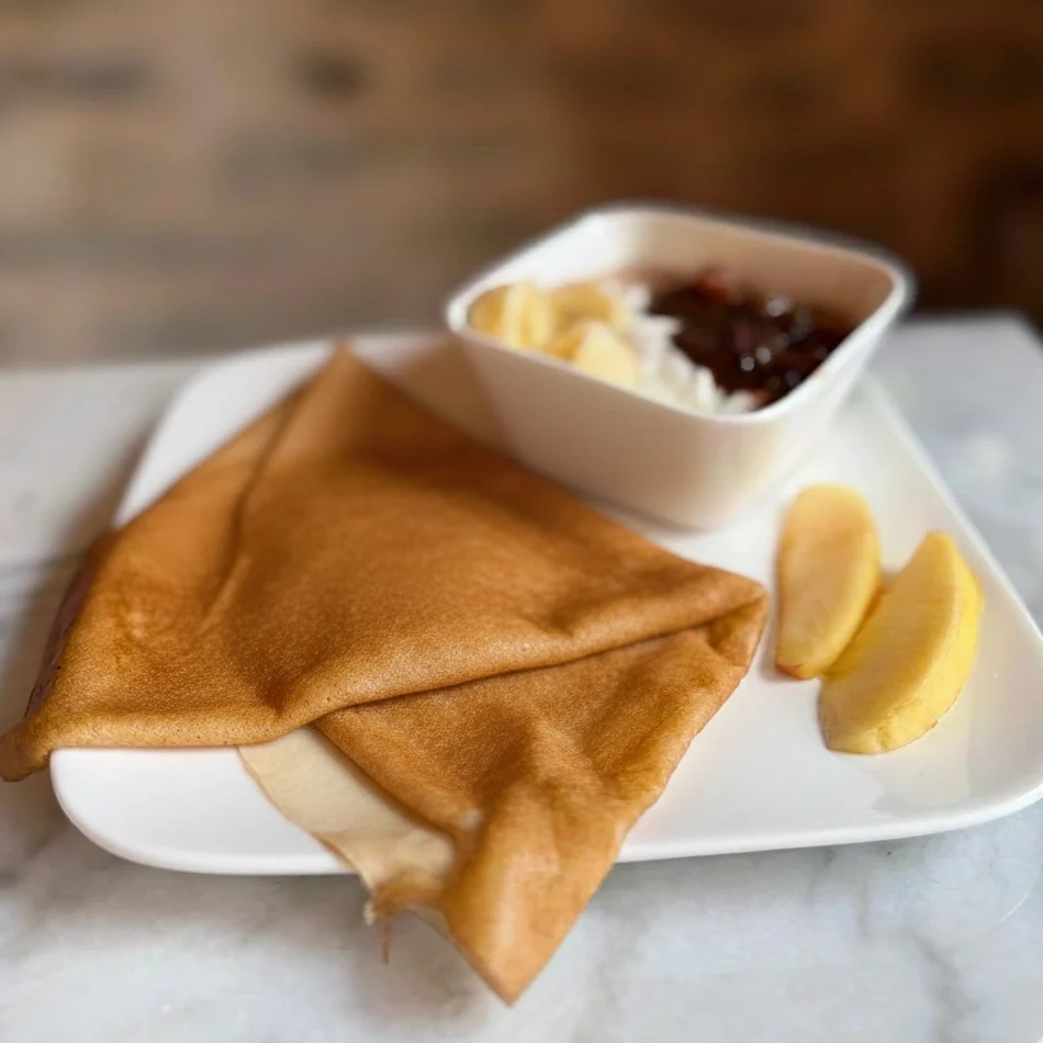 Une crêpe dorée pliée en triangle, posée sur une assiette en porcelaine blanche décorée de baies, sur une table en bois texturé.